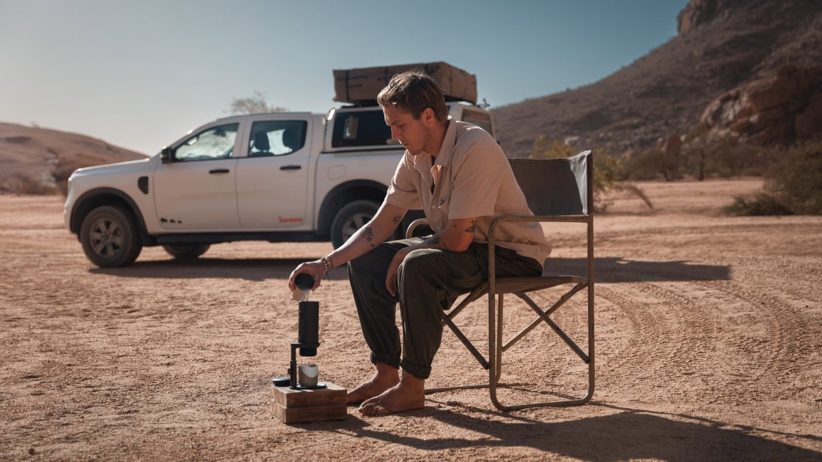 A man in a camp chair in the desert pouring water into a portable espresso maker, a pickup truck behind him